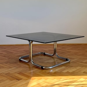 May include: A square coffee table featuring a dark smoked glass top supported by a polished chrome tubular metal frame. The base consists of a continuous bent metal structure with black connectors at the joints, providing a stable foundation.