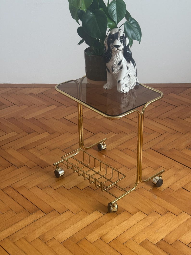 May include: A gold metal rolling cart with a glass top and a wire basket underneath. The cart has four wheels and is sitting on a wooden floor. A black and white porcelain dog figurine is sitting on the glass top.