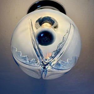 May include: A clear glass globe light fitting with a decorative, flowing design. The fitting has a black base and a visible lightbulb inside. The glass has a textured, rippled appearance, creating a unique aesthetic.