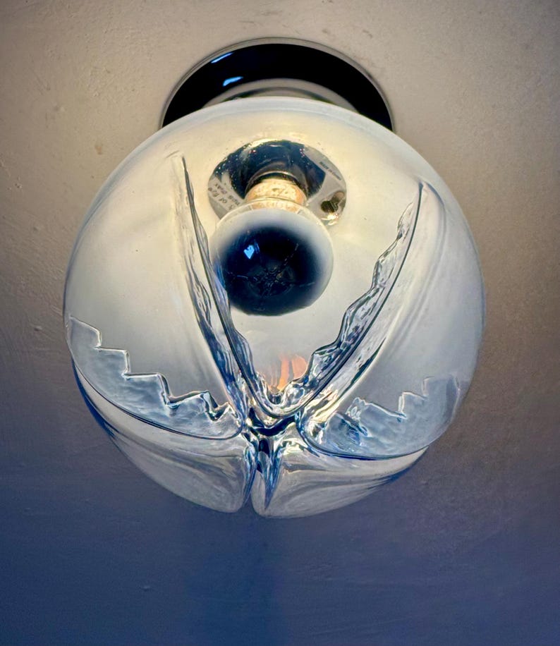 May include: A clear glass globe light fitting with a decorative, flowing design. The fitting has a black base and a visible lightbulb inside. The glass has a textured, rippled appearance, creating a unique aesthetic.