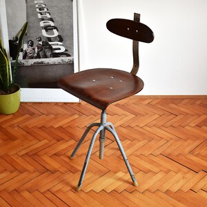 Puede incluir: Una silla giratoria industrial vintage con un asiento de madera marr&oacute;n y una base de metal gris. La silla tiene un respaldo de metal negro con una parte superior curva.