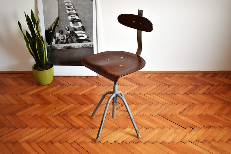 Puede incluir: Una silla giratoria industrial vintage con un asiento de madera marr&oacute;n y una base de metal gris. La silla tiene un respaldo de metal negro con una parte superior curva.
