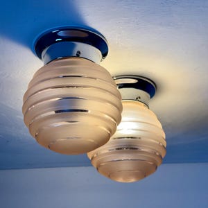 May include: Two ceiling-mounted light fixtures. Each fixture has a chrome base and a frosted glass shade with horizontal ridges. The shades are a soft, warm color, and the fixtures are set against a blue ceiling.