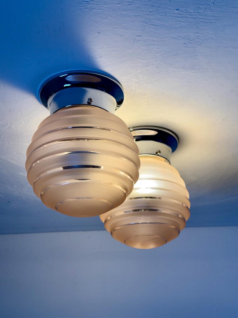 May include: Two ceiling-mounted light fixtures. Each fixture has a chrome base and a frosted glass shade with horizontal ridges. The shades are a soft, warm color, and the fixtures are set against a blue ceiling.