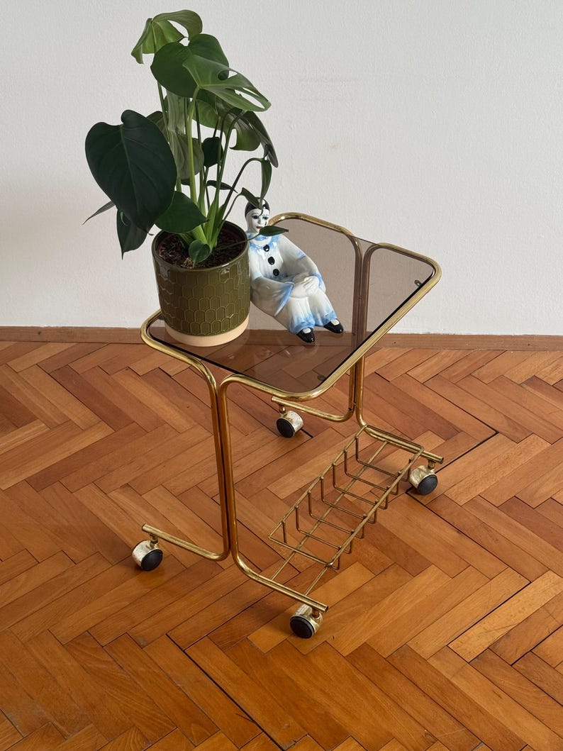 May include: A gold metal rolling cart with a glass top and a wire shelf. A green potted plant and a porcelain figurine are on the top shelf. The cart is on a wooden floor.