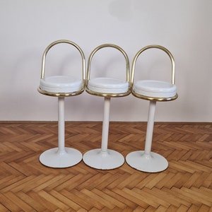 May include: Three white and gold bar stools with a rounded seat and a curved back. The stools are arranged in a row on a wooden floor.