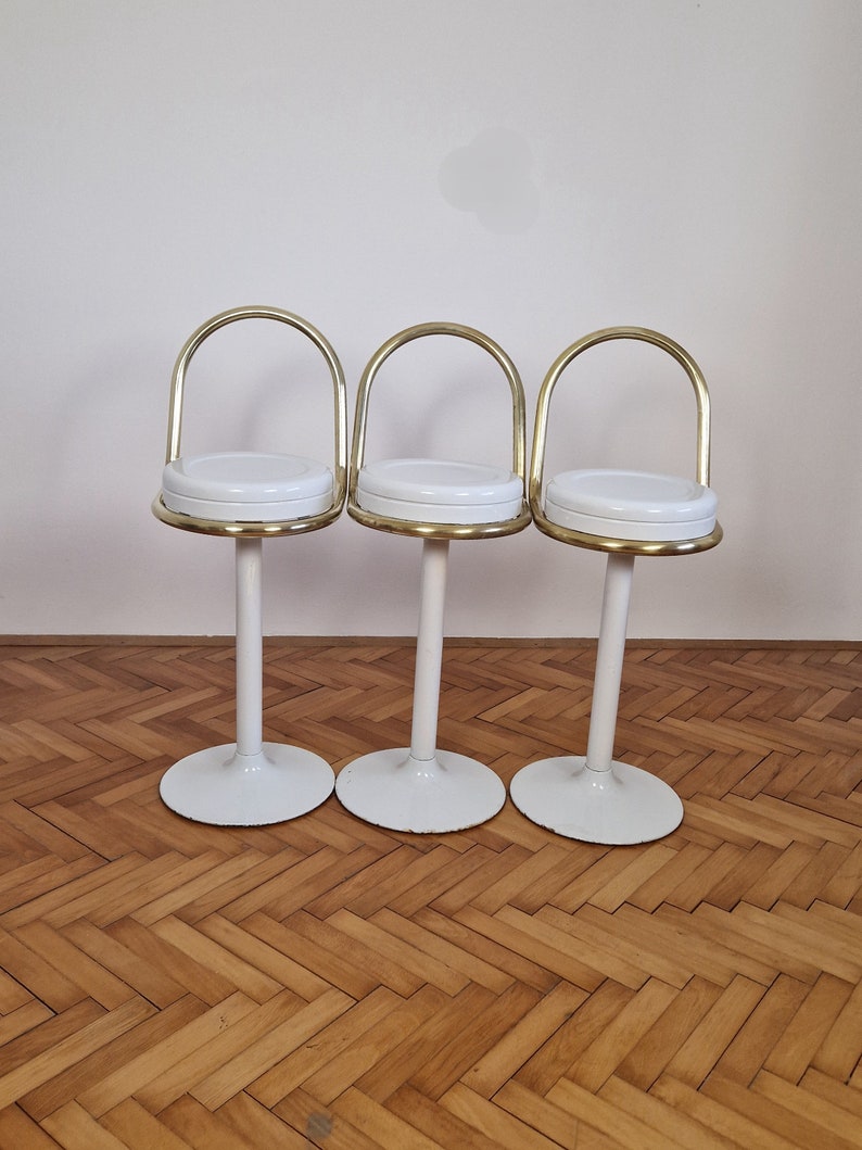 May include: Three white and gold bar stools with a rounded seat and a curved back. The stools are arranged in a row on a wooden floor.