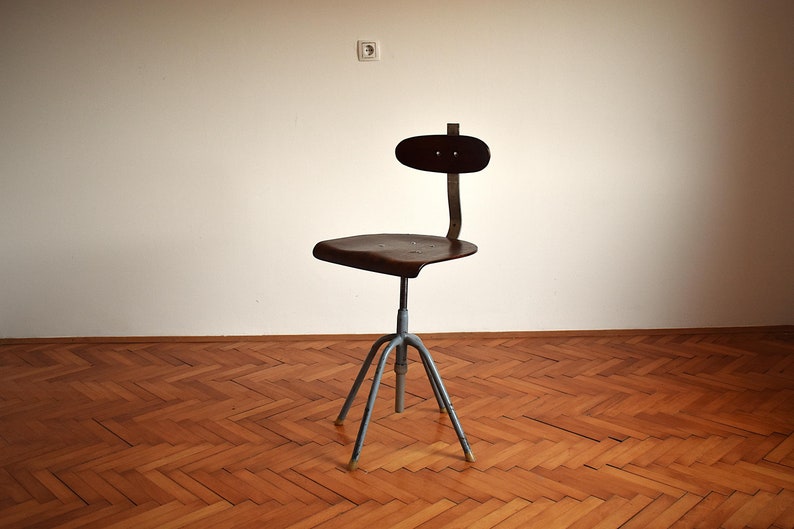 Puede incluir: Una silla giratoria industrial vintage con asiento y respaldo de madera marr&oacute;n oscuro. La silla tiene una base de metal con cuatro patas y una palanca de ajuste de altura.