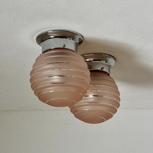 May include: Two Art Deco-style ceiling lights. Each light has a chrome base and a frosted, peach-colored glass shade with horizontal ridges. The lights are mounted on a white ceiling.