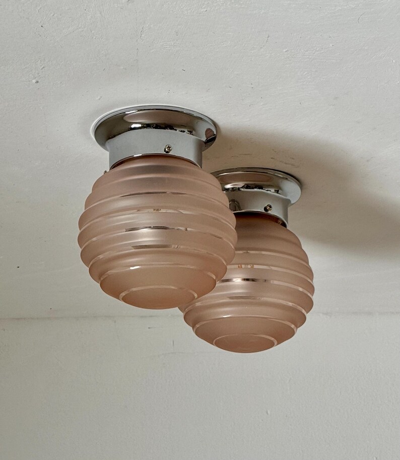 May include: Two Art Deco-style ceiling lights. Each light has a chrome base and a frosted, peach-colored glass shade with horizontal ridges. The lights are mounted on a white ceiling.