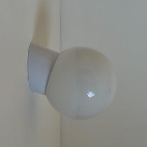 May include: A white, spherical wall-mounted light fixture. The fixture has a rounded, opaque glass globe and a white, angled base attached to the wall. The light is designed to be mounted in a corner.