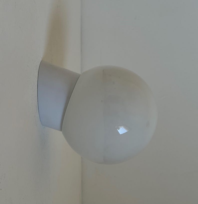 May include: A white, spherical wall-mounted light fixture. The fixture has a rounded, opaque glass globe and a white, angled base attached to the wall. The light is designed to be mounted in a corner.