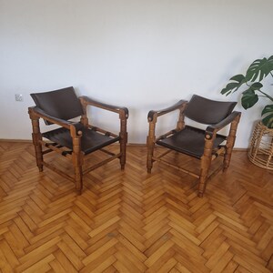 May include: Two brown leather chairs with wooden frames. The chairs have a simple design with a rectangular seat and back. The chairs are sitting on a wooden floor with a herringbone pattern.
