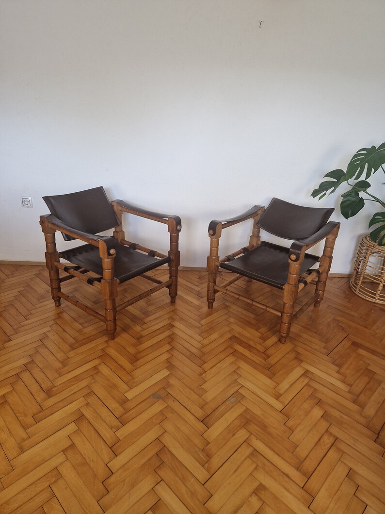 May include: Two brown leather chairs with wooden frames. The chairs have a simple design with a rectangular seat and back. The chairs are sitting on a wooden floor with a herringbone pattern.