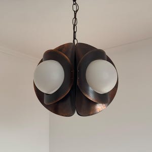 Puede incluir: Una l&aacute;mpara colgante de tono cobre con una cadena que cuelga del techo. La l&aacute;mpara tiene un dise&ntilde;o geom&eacute;trico con dos globos de vidrio blanco esmerilado.