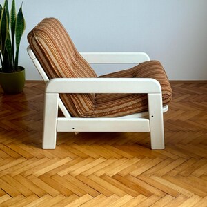 May include: A white wooden armchair with a brown and beige striped fabric cushion. The chair is sitting on a wooden floor with a herringbone pattern.