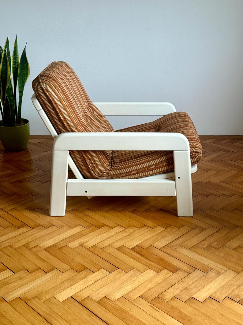 May include: A white wooden armchair with a brown and beige striped fabric cushion. The chair is sitting on a wooden floor with a herringbone pattern.