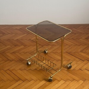 May include: A gold metal side table with a smoked glass top and a wire basket underneath. The table has four wheels and is on a wooden floor.