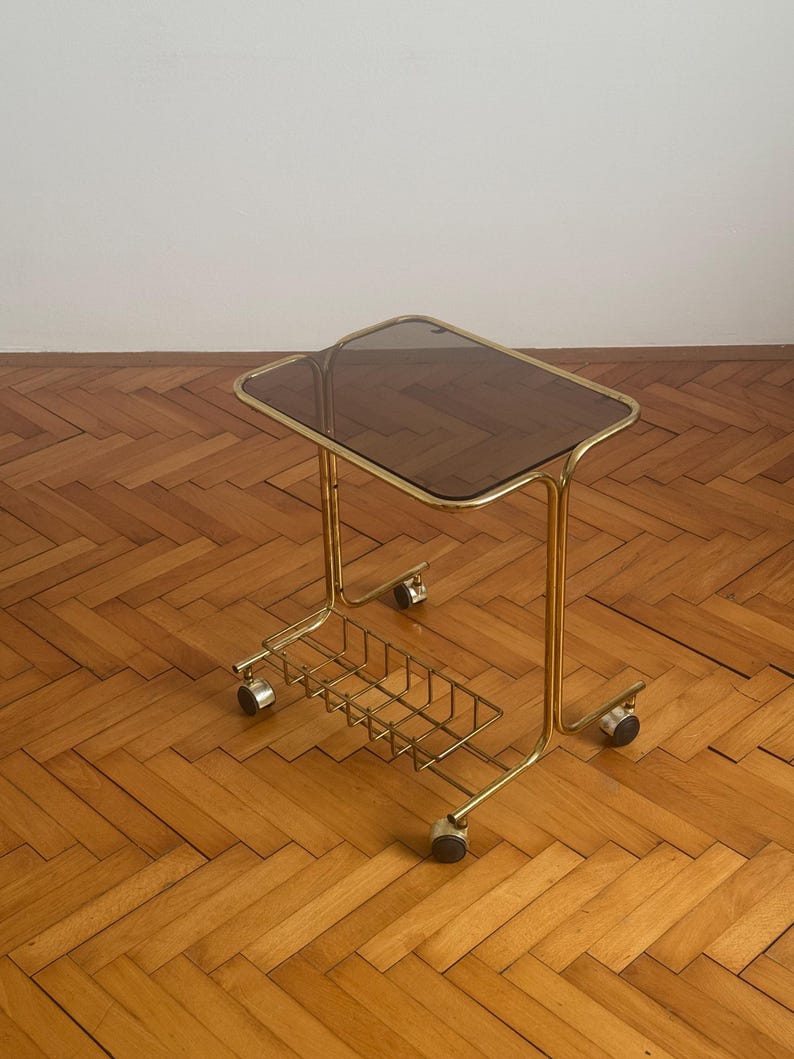May include: A gold metal side table with a smoked glass top and a wire basket underneath. The table has four wheels and is on a wooden floor.