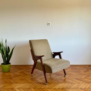 May include: A mid-century modern armchair with a beige fabric upholstery and dark wood legs. The chair is on a wood floor with a herringbone pattern.