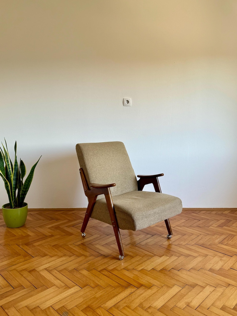 May include: A mid-century modern armchair with a beige fabric upholstery and dark wood legs. The chair is on a wood floor with a herringbone pattern.