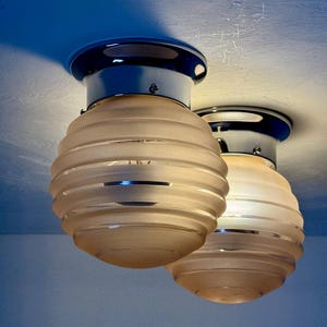 May include: Two ceiling-mounted light fixtures. Each fixture has a chrome base and a frosted glass shade with horizontal ridges. The shades are a warm, soft white color, and the chrome reflects the light. The fixtures are mounted on a light blue ceiling.