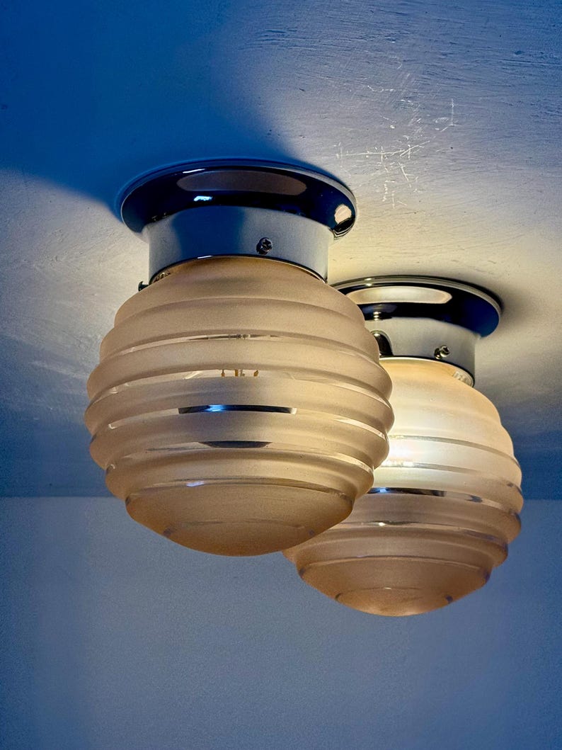 May include: Two ceiling-mounted light fixtures. Each fixture has a chrome base and a frosted glass shade with horizontal ridges. The shades are a warm, soft white color, and the chrome reflects the light. The fixtures are mounted on a light blue ceiling.