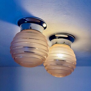 May include: Two Art Deco-style ceiling lights. Each light has a chrome base and a frosted, ribbed glass shade. The shades are a soft peach color, and the lights are mounted on a white ceiling.
