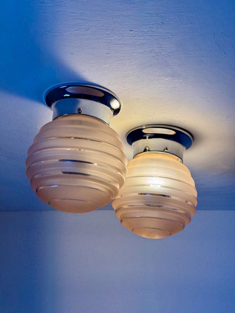 May include: Two Art Deco-style ceiling lights. Each light has a chrome base and a frosted, ribbed glass shade. The shades are a soft peach color, and the lights are mounted on a white ceiling.