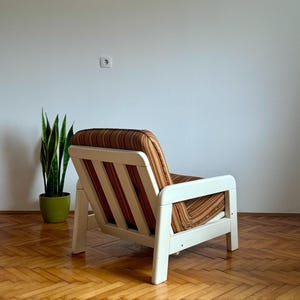 May include: A white wooden armchair with a brown and orange striped fabric upholstery. The chair is sitting on a wooden floor with a herringbone pattern.