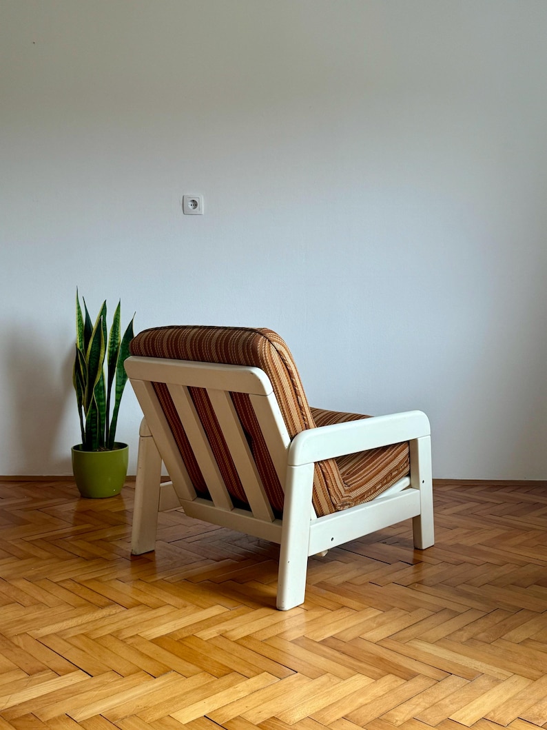 May include: A white wooden armchair with a brown and orange striped fabric upholstery. The chair is sitting on a wooden floor with a herringbone pattern.