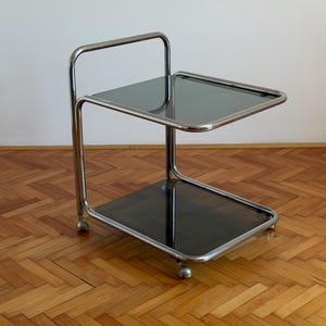 Puede incluir: Una mesa auxiliar con ruedas de dos niveles que presenta un marco tubular de metal cromado pulido y dos estantes cuadrados de vidrio ahumado oscuro. La estructura incluye un asa alta en un lado y cuatro pequeñas ruedas.