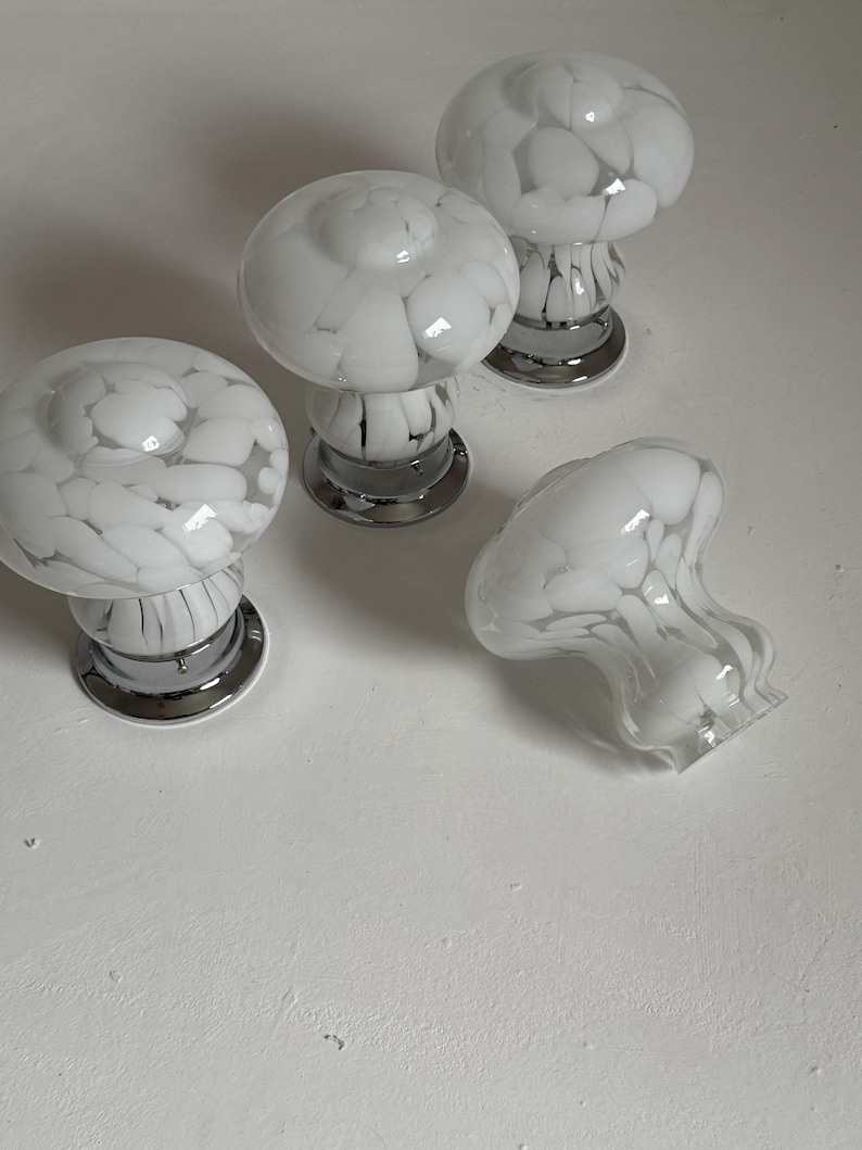 May include: Four white and gray glass mushroom-shaped cabinet knobs with chrome bases. One knob is lying on its side. The knobs have a speckled pattern and are designed for furniture or decorative use.