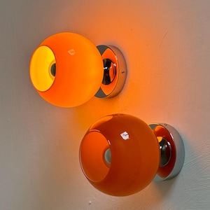 May include: Two orange globe-shaped wall sconces with a chrome base. The sconces have a circular design with a cut-out on one side, revealing a yellow interior. The light fixtures are mounted on a white wall.