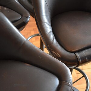 Op de afbeelding: Vier bruine leren barstoelen met een metalen basis. De krukken zijn in een rij geplaatst, met de zitting van elke kruk naar de camera gericht.