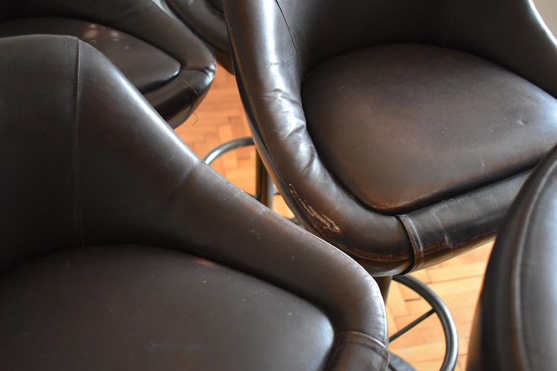 Op de afbeelding: Vier bruine leren barstoelen met een metalen basis. De krukken zijn in een rij geplaatst, met de zitting van elke kruk naar de camera gericht.