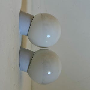 May include: Two white globe-shaped wall sconces with gray mounting brackets are mounted on a light-colored wall. The sconces are positioned one above the other, creating a symmetrical design. The globes have a glossy finish.