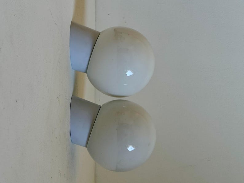 May include: Two white globe-shaped wall sconces with gray mounting brackets are mounted on a light-colored wall. The sconces are positioned one above the other, creating a symmetrical design. The globes have a glossy finish.