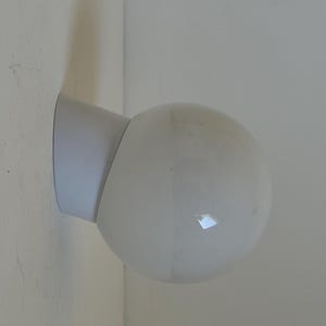 May include: A white, spherical wall-mounted light fixture. The fixture has a rounded, globe-shaped light and a cylindrical arm that attaches to a flat, rectangular base. The light is mounted on a white wall.
