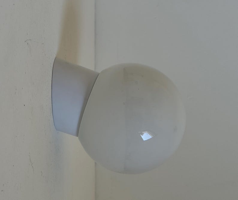 May include: A white, spherical wall-mounted light fixture. The fixture has a rounded, globe-shaped light and a cylindrical arm that attaches to a flat, rectangular base. The light is mounted on a white wall.