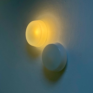 May include: Two circular wall-mounted light fixtures with smooth, rounded profiles. One fixture emits a warm, glowing yellow light, while the other is opaque white and unlit. Both fixtures feature a simple, clean design mounted on a flat surface.