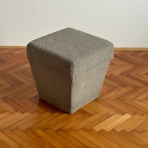 May include: A square, upholstered ottoman with a striped pattern in shades of gray and beige. The ottoman has a trapezoidal shape, wider at the top and narrower at the base. It sits on a wooden floor.