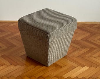 Może przedstawiać: Kwadratowy, tapicerowany puf z wzorem w paski w odcieniach szarości i beżu. Puf ma trapezoidalny kształt, szerszy u góry i węższy u podstawy. Stoi na drewnianej podłodze.