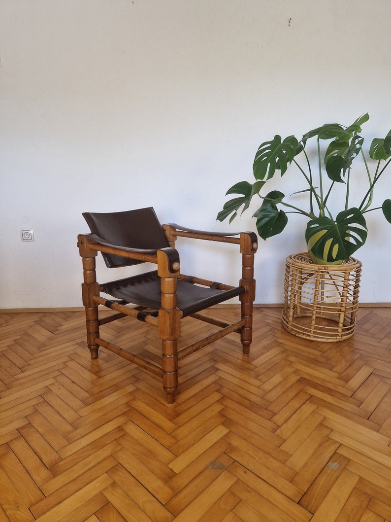 May include: A brown wooden chair with a black leather seat and back. The chair has a simple design with four legs and a rectangular frame. The chair is sitting on a wooden floor with a herringbone pattern.