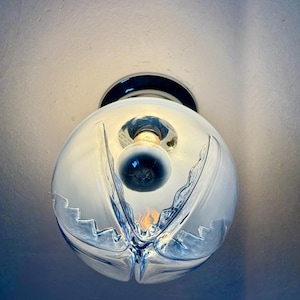 May include: A clear glass globe light fitting with a decorative, wave-like design. The light bulb is visible inside the globe. The fitting is mounted on a dark, circular base, and is attached to a white ceiling.