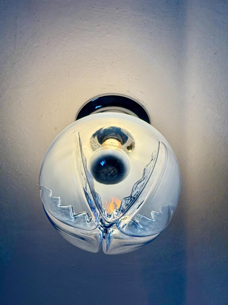 May include: A clear glass globe light fitting with a decorative, wave-like design. The light bulb is visible inside the globe. The fitting is mounted on a dark, circular base, and is attached to a white ceiling.
