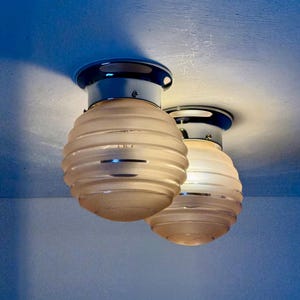 May include: Two ceiling-mounted light fixtures. Each fixture has a chrome base with a dark blue rim and a frosted glass shade. The shades are spherical with horizontal ribbed detailing. The lights are against a blue background.