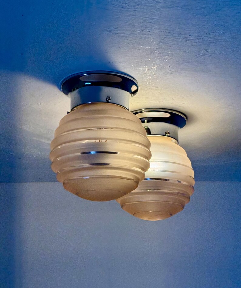 May include: Two ceiling-mounted light fixtures. Each fixture has a chrome base with a dark blue rim and a frosted glass shade. The shades are spherical with horizontal ribbed detailing. The lights are against a blue background.