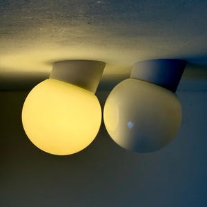 May include: Two modern, spherical ceiling lights. One is illuminated with a warm yellow glow, while the other is unlit. Each light has a white base and a rounded, off-white globe. The lights are mounted on a light-colored ceiling.