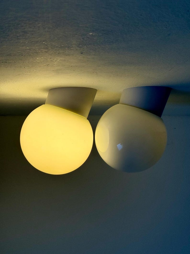May include: Two modern, spherical ceiling lights. One is illuminated with a warm yellow glow, while the other is unlit. Each light has a white base and a rounded, off-white globe. The lights are mounted on a light-colored ceiling.